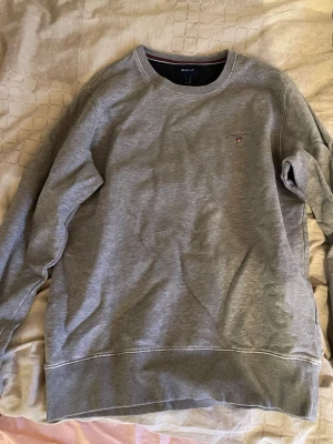 Grå sweatshirt från GANT - Säljer en stilren grå sweatshirt från GANT med rund hals och diskret logga broderad på bröstet. Tröjan har långa ärmar och ribbade muddar vid ärmslut och nederkant. Perfekt för en chill och avslappnad look.