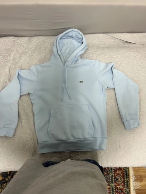 Lacoste Hoodie - Den här ljusblå Lacoste-hoodien kombinerar tidlös stil med premiumkomfort i ett mjukt och slitstarkt material. Den ikoniska krokodilloggan och den perfekta passformen gör den lika snygg till vardags som till helgen. För 999 kr får du en hoodie som håller länge och alltid ser rätt ut.