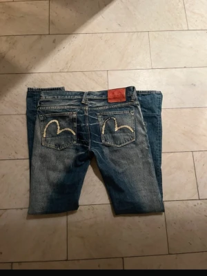 Evisu Genes blå jeans med broderi - Säljer ett par klassiska blå jeans från Evisu Genes med ikoniskt vitt märke på bakfickorna och röd läderpatch i midjan. Jeansen har knäppning framtill och slitna detaljer som ger en cool vintagekänsla. Passformen är slim/straight 