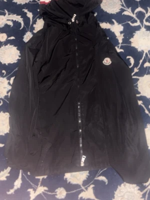 Svart vindjacka från Moncler - Svart vindjacka från Moncler med huva och dragkedja framtill. Jackan har Moncler-logga på bröstet och en snygg randig detalj i rött, vitt och blått på huvan. Perfekt för dig som vill ha en stilren och sportig look.