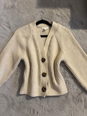 Beige ribbad kofta  - Snygg beige kofta med ribbad struktur och tre stora bruna knappar framtill. Modellen har v-ringad hals och lång ärm. Materialet är stickat och ger en mjuk och skön känsla. Använd fåtal gånger, mycket bra skick. Inget att anmärka 