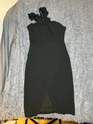 Svart one shoulder-klänning med volang - Snygg svart klänning med omlottkjol och volangdetaljer vid halsen. Materialet känns som polyester och passformen är normal. Perfekt för dig som gillar stilrena och trendiga plagg.