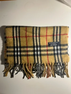 Rutig Burberry cashmere halsduk - Säljer en klassisk rutig halsduk från Burberry i 100% cashmere. Färgerna är beige, svart, vit och röd med fransar i kanterna. Mjuk och lyxig känsla, tillverkad i England. Perfekt för att lyfta din stil med ikoniskt mönster.