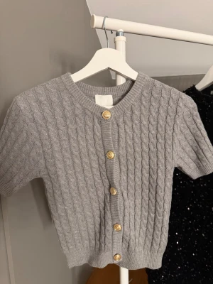 Grå kabelstickad kofta från H&M - Säljer en grå kabelstickad kofta från H&M i storlek XS. Koftan har korta ärmar, rund hals och snygga guldfärgade knappar framtill. Prislapp kvar då den aldrig är använd🤍
