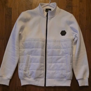 Vit quiltad jacka Philipp Plein - Snygg vit jacka från Philipp Plein i storlek 2XL men sitter som L/XLmed quiltade partier och svart dragkedja. Jackan har ett svart PP-märke på bröstet och en stor broderad döskalle med logga på ryggen. Perfekt för dig som vill sticka ut med en unik och modern look.