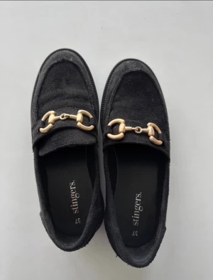 Svarta loafers från Stingers, strl 37 - Säljer ett par svarta loafers från Stingers i storlek 37. Skorna har en snygg guldfärgad metall-detalj över foten och är gjorda i mocka med platt sula och rund tå.