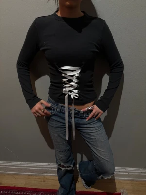 Occasio - Säljer en svart långärmad topp med vit snörning framtill som ger en korsettkänsla. Toppen är croppad och har en tight passform. Materialet är ribbat och stretchigt, vilket gör den både bekväm och snygg. Perfekt för dig som vill ha en edgy och trendig look. (Copenhagen) 🌟i storlek S