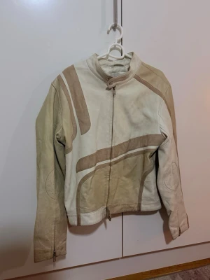 Beige skinnjacka Deadwood M - Säljer en beige och ljusbrun skinnjacka från Deadwood i storlek M. Jackan har coola grafiska paneler i olika nyanser, ståkrage med spänne och dragkedja framtill. Materialet är mjukt skinn och jackan har långa ärmar med dragkedja vid ärmslutet.