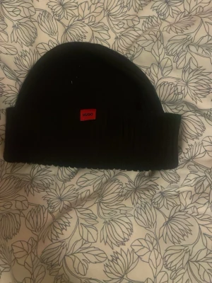 Svart mössa från Hugo - Svart ribbstickad mössa från Hugo med uppvikt kant och en liten röd logotyp framtill. Enkel och stilren design som passar till de flesta outfits. Mjuk och skön, perfekt för kyliga dagar.