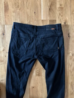 Svarta jeansbyxor från BOSS 34/32 - Svarta jeansbyxor från BOSS med klassisk femficksdesign och snygga detaljer i orange. Byxorna har rak passform och är tillverkade i mjuk bomull med lite stretch för extra komfort. Perfekt för dig som gillar stilrena och tidlösa plagg.