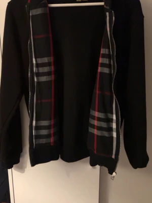 Svart zip tröja från Burberry  - Svart zip tröja från Burberry med klassiskt rutigt foder i rött, grått och vitt. Tröjan  har dragkedja framtill och ribbade muddar. Tillverkad i bomull och har en clean, stilren look med ikoniska Burberry-detaljer på insidan. Använd endast 4-5 gånger, inga skador. Materialet är i nästan nyskick