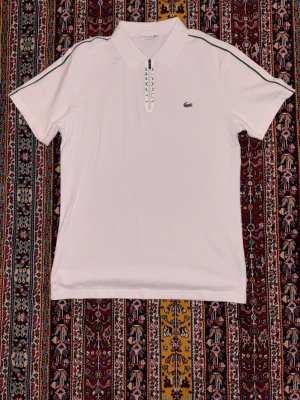 Lacoste piké - Vit polotopp från Lacoste i klassisk design med korta ärmar och grön logotyp på bröstet. Dragkedja framtill och gröna ränder längs axlarna ger en sportig vibe. Tillverkad i bomull, perfekt för dig som gillar stilren och clean look.