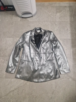 Silver kavaj från Kappahl, storlek 42 - Säljer en riktigt snygg silverfärgad kavaj från Kappahl i storlek 42. Kavajen har dubbelknäppning, klassisk krage och två fickor fram. Materialet är glansigt och har en cool metallic-look som verkligen sticker ut. Perfekt för dig som vill ha något unikt i garderoben. Använd en gång, som ny
