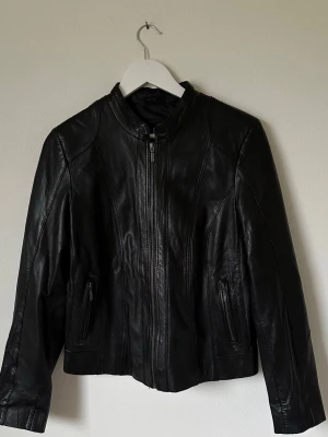 Svart skinnjacka från SAKI, strl 40 - Snygg svart skinnjacka från SAKI i äkta skinn med stilren design och raka linjer. Jackan har ståkrage, dragkedja framtill och två fickor med dragkedja. Perfekt för dig som vill ha en cool och tidlös look. 7Y4