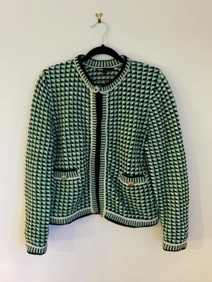 Rutig cardigan jacka från Maje - Snygg cardigan jacka från Maje med rutigt mönster i grönt, svart och vitt. Jackan har rund krage, två fickor med guldfärgade knappar och stickade detaljer längs kanterna. Perfekt för dig som gillar statement-plagg och coola mönster.
