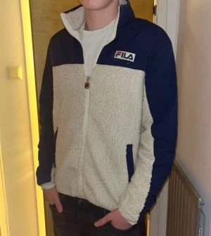 FILA teddyjacka  - Säljer en beige och marinblå teddyjacka från FILA med dragkedja och högt krage. Jackan har två fickor med marinblå detaljer och ett broderat FILA-märke på bröstet. Materialet är mjuk polyester och jackan är långärmad med normal passform. Axelbredd 45cm, längd 68cm. OBS finns ingen storlekslapp men uppskattas till en medium. JAG SKICKAR INGA FLER BILDER NÄR JAG BÄR PLAGGET PÅ :) !!
