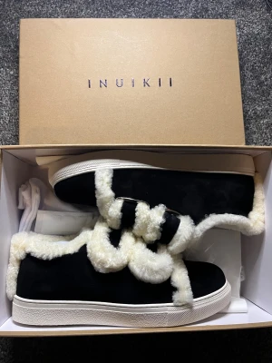 Svarta sneakers från Inuikii med päls - Säljer ett par svarta sneakers från Inuikii med fluffig vit pälsdetalj runt kanten och på snörningen. Skorna har en platt vit sula och är tillverkade i mocka med päls för extra mysig känsla. Perfekta för kyliga dagar och riktigt snygga till jeans. Storlek 39!