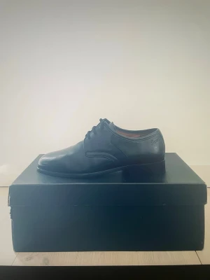 Svarta skinnskor från Sioux - Klassiska svarta skinnskor från Sioux, modell Rochester. Snygg rund tå och platt sula, med stilren snörning och diskret logga på sidan. Tillverkade i mjukt lammnappa och har lädersula för extra komfort. Perfekt för dig som gillar tidlös och clean stil.