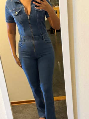 Blå jeans jumpsuit med dragkedja - Säljer en snygg blå jeans jumpsuit med korta ärmar och krage. Jumpsuiten har en framträdande dragkedja hela vägen upp, fickor fram och bak samt bälteshällor. Materialet är stretchigt jeans och passformen är tight med raka ben. Perfekt för dig som gillar en cool och modern look.