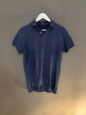 Mörkblå piké från Polo Ralph Lauren - Säljer en mörkblå pikétröja från Polo Ralph Lauren i storlek M. Tröjan har klassisk krage, två knappar vid halsen och den ikoniska röda broderade loggan på bröstet. Materialet är mjukt och känns som bomull, perfekt för en chill och stilren look.