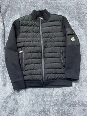 Svart pufferjacka från Moncler S - Svart pufferjacka från Moncler i storlek S med quiltad framsida och släta ärmar. Jackan har dragkedja framtill, två fickor med dragkedja och en ficka med Moncler-logga på ärmen. Materialet är polyester och syntet, perfekt för kyliga dagar.