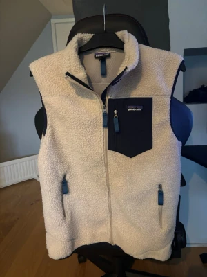 Patagonia fleeceväst  - Säljer en beige fleeceväst från Patagonia med marinblå detaljer. Västen har hög krage, tre fickor med dragkedja och ett mjukt, fluffigt material som håller dig varm. Perfekt att slänga över en hoodie eller tröja när det är kyligt ute. Skick 9/10. Hör av er vid frågor 