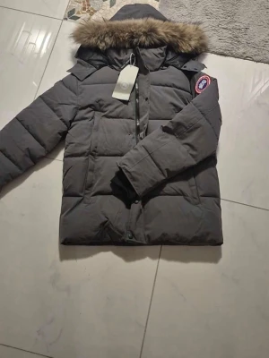 Mörkgrå dunjacka från Canada Goose - Säljer en mörkgrå dunjacka från Canada Goose med pälskantad huva och klassisk logga på ärmen. Jackan har långärmade quiltade partier, dragkedja och knappar framtill. Insidan är fodrad med svart satin för extra komfort och värme.
