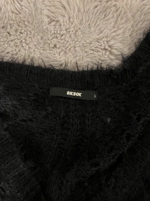 Svart stickad Bik Bok tröja v-ringad - Mysig svart stickad tröja från Bik Bok i storlek S. Tröjan har ett fluffigt material, v-ringad hals och ett snyggt hålmönster både fram och bak.