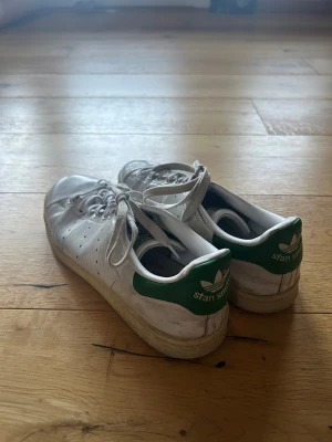 Adidas Stan Smith vita sneakers - Säljer ett par klassiska Adidas Stan Smith sneakers i vitt med gröna detaljer på hälen och tungan. Skorna har snörning, rund tå och platt sula. De är använda och skavankerna syns på bild. Snygga att styla till ett par bredare jeans. 