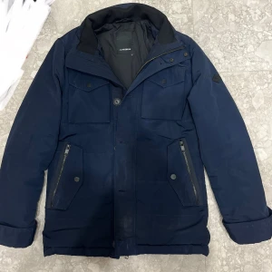 field jacket j.lindeberg - xs passar s också Säljer en mörkblå pufferjacka från J.Lindeberg med flera fickor, dragkedja och knappar framtill. Jackan har en hög krage och snyggt JL-märke på ärmen. Materialet är 100% polyester och jackan är perfekt för kalla dagar.