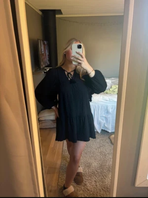 Svart kort klänning från Gina Tricot - Supersöt svart kort klänning från Gina Tricot med långa ärmar och knytning vid halsen. Klänningen har en lätt strukturerad yta och en lös passform som ger en chill vibe. Perfekt för dig som gillar enkel men snygg stil.