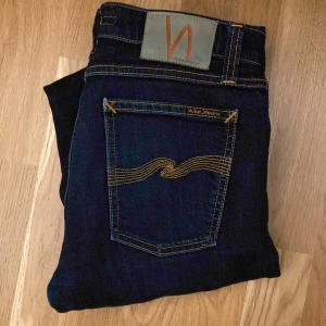 Nudie jeans grim Tim  - Säljer ett par mörkblå jeans från Nudie Jeans med klassisk femficksdesign och snygga kontrastsömmar. Jeansen har en rak passform och är tillverkade i slitstarkt denim med Nudie Jeans logga på bakfickan. Perfekt för dig som gillar stilrena och tidlösa jeans.