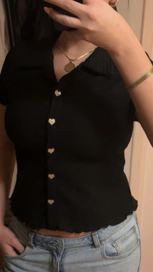 Svart blus med hjärtknappar - Säljer en svart blus med korta ärmar och krage. Blusen har söta hjärtformade knappar framtill och är lätt ribbad i materialet. Den är figurnära och har vågig kant nedtill och vid ärmslut. Perfekt att styla med jeans eller kjol.