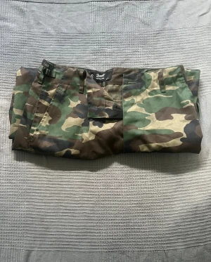 Cargopants i camouflage från Brandit - Säljer ett par cargopants från Brandit med klassiskt camouflage-mönster i grönt, brunt och beige. Byxorna har flera praktiska fickor på sidorna och baktill, samt justerbara spännen i midjan. Perfekta för dig som gillar streetwear och utility-style.