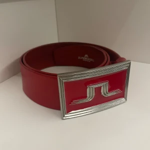 J.Lindberg Stockholm Skärp - Ett rött unikt skärp från J.Lindberg. | Skärpet är cirka 100cm långt. | Det är köpt secondhand och har lite tecken på användning, så som små repor och tryck märken. | Det är bara att höra av sig om man har några frågor eller om man vill ha flera bilder.