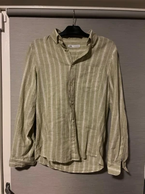 Randig grön skjorta från Zara - Snygg grön skjorta från Zara med vita vertikala ränder. Skjortan har klassisk krage, bröstficka och knappar framtill. Tillverkad i linne, vilket gör den luftig och skön. Perfekt för dig som gillar en chill och stilren look.