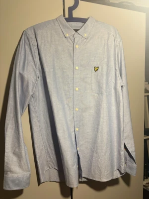 Ljusblå skjorta från Lyle & Scott - Snygg ljusblå skjorta från Lyle & Scott med klassisk krage och gul fågel-logga på bröstfickan. Skjortan har vita knappar framtill och är långärmad. Perfekt för dig som gillar stilrena och enkla plagg med en touch av märke. Skjortan är i nytt skick!