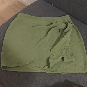 Grön omlottkjol med knyt - Snygg grön omlottkjol med knyt i midjan och diskret dragkedja bak. Kjolen har en kort längd och är gjord i ett lätt, krinklat tyg som ger en avslappnad vibe. Perfekt för dig som gillar stilrena plagg med en twist. Passar till sneakers eller sandaler.