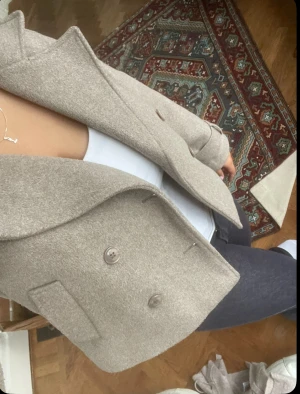 Beige kappa från Gina Tricot - Säljer en beige kavaj från Gina Tricot i storlek M. Kavajen har dubbelknäppning med stora knappar, klassisk krage och är tillverkad i ett mjukt ull-liknande material. Perfekt att styla med jeans eller kjol för en snygg look. Skriv gärna för fler/tydliga bilder 