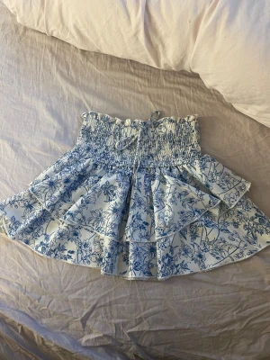 Blommig volangkjol med snörning - Supersöt kort kjol med blått blommönster på vit botten. Kjolen har bred smock i midjan och snörning framtill, samt flera lager volanger som ger en härlig och lekfull look. Perfekt för dig som gillar romantiska och fräscha vibes.