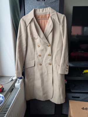 Beige kappa med knappar - Snygg beige kappa med dubbelknäppning och stora knappar framtill och vid ärmslut. Klassisk krage och fickor med lock ger en stilren look. Materialet känns som bomull och kappan har en mjuk, ljus ton. Perfekt för dig som gillar tidlös och clean stil.
