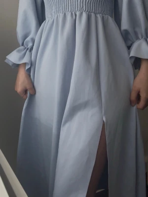 Ljusblå långklänning med slits - Säljer en ljusblå långklänning med smock upptill och vida ärmar med knyt vid handlederna. Klänningen har en snygg slits framtill som ger extra edge. Materialet känns mjukt och flowy, perfekt för dig som gillar stilrena plagg med coola detaljer.