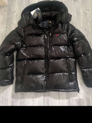 Svart pufferjacka från Polo Ralph Lauren - Säljer en svart pufferjacka från Polo Ralph Lauren med glansig finish och röd broderad logga på bröstet. Jackan har huva, dragkedjor framtill och sidofickor. Perfekt för kalla dagar och har en riktigt schysst streetstil.