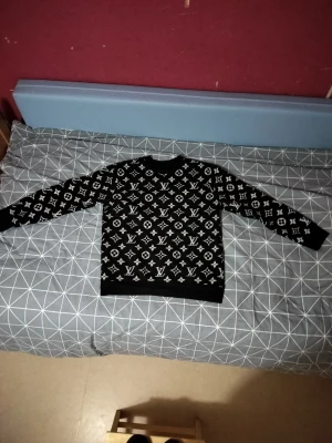 Svart Louis Vuitton tröja  - Svart tröja från Louis Vuitton med ikoniskt monogrammönster i vitt över hela tröjan. Rund hals och långa ärmar. Mjuk och bekväm, perfekt för dig som gillar exklusiv streetwear. Märkesetikett i nacken och insida med QR-kod. Snygg och stilren design.