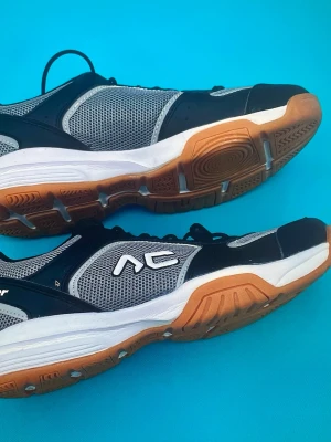 Svarta och grå sneakers från AC, strl 39 - Säljer ett par snygga sneakers från AC i storlek 39. Skorna har svart och grå mesh med vita detaljer och en gummisula i brunt. De har snörning och en sportig design med rund tå och platt sula, perfekta för träning eller häng.