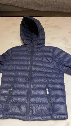 Blå dunjacka Polo Ralph Lauren XL barn  - Säljer en mörkblå dunjacka från Polo Ralph Lauren i storlek XL barn. Jackan har huva, två fickor med dragkedja och quiltad design. Perfekt för kalla dagar och har en stilren look med diskret logga på bröstet, det finns ett litet hål på ryggen se bild 4 