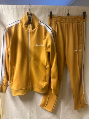 Palm Angels senapsgul tracksuit - Säljer en snygg senapsgul tracksuit från Palm Angels med vita och svarta ränder längs ärmar och ben. Setet består av zip-jacka med hög krage och två fickor med dragkedja samt matchande byxor med elastisk midja. Palm Angels-logga i vitt på både jacka och byxa. Helt ny. 