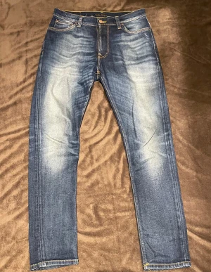 Blå Nudie Jeans Brute Knut W29 L30 - Snygga blå jeans från Nudie Jeans, modellen Brute Knut. De har klassisk femficksdesign, orangea sömmar och coola slitningar framtill och baktill. Materialet är 99% ekologisk bomull och 1% elastan för extra komfort. Passformen är rak med något smalare ben. Endast använda vid 2 tillfällen.  Bara att höra av sig vid minsta fundering. Pris kan diskuteras. 
