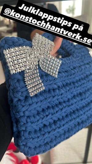 Blå virkad  handväska med rosett - Unik blå stickad handväska från Konst och Hantverk, dekorerad med en stor glittrig rosett i silver. Väskan har ett handtag upptill och är perfekt för dig som gillar personliga accessoarer med extra bling. Handgjord och riktigt snygg att bära till din outfit.
