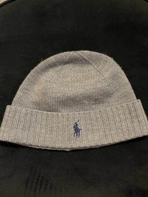 Grå mössa från Polo Ralph Lauren - Säljer en stilren grå mössa från Polo Ralph Lauren i mjuk merinoull. Mössan har ribbad kant och klassisk Polo-logga broderad framtill i mörkblått. Perfekt för kalla dagar och enkel att matcha med din jacka. Pris går att diskutera 